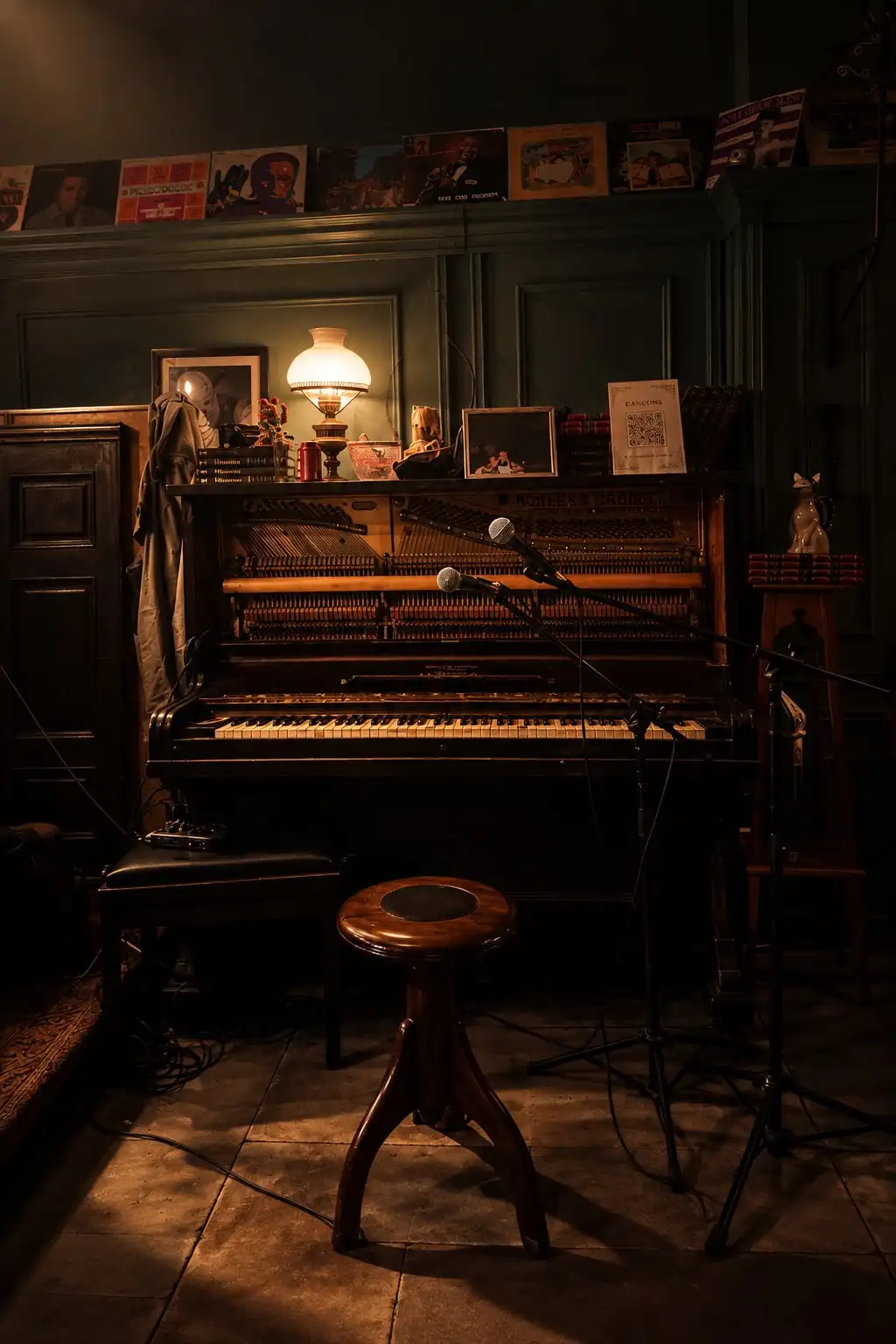 Un piano vertical antiguo con un taburete de madera, una lámpara encendida y discos en la pared.