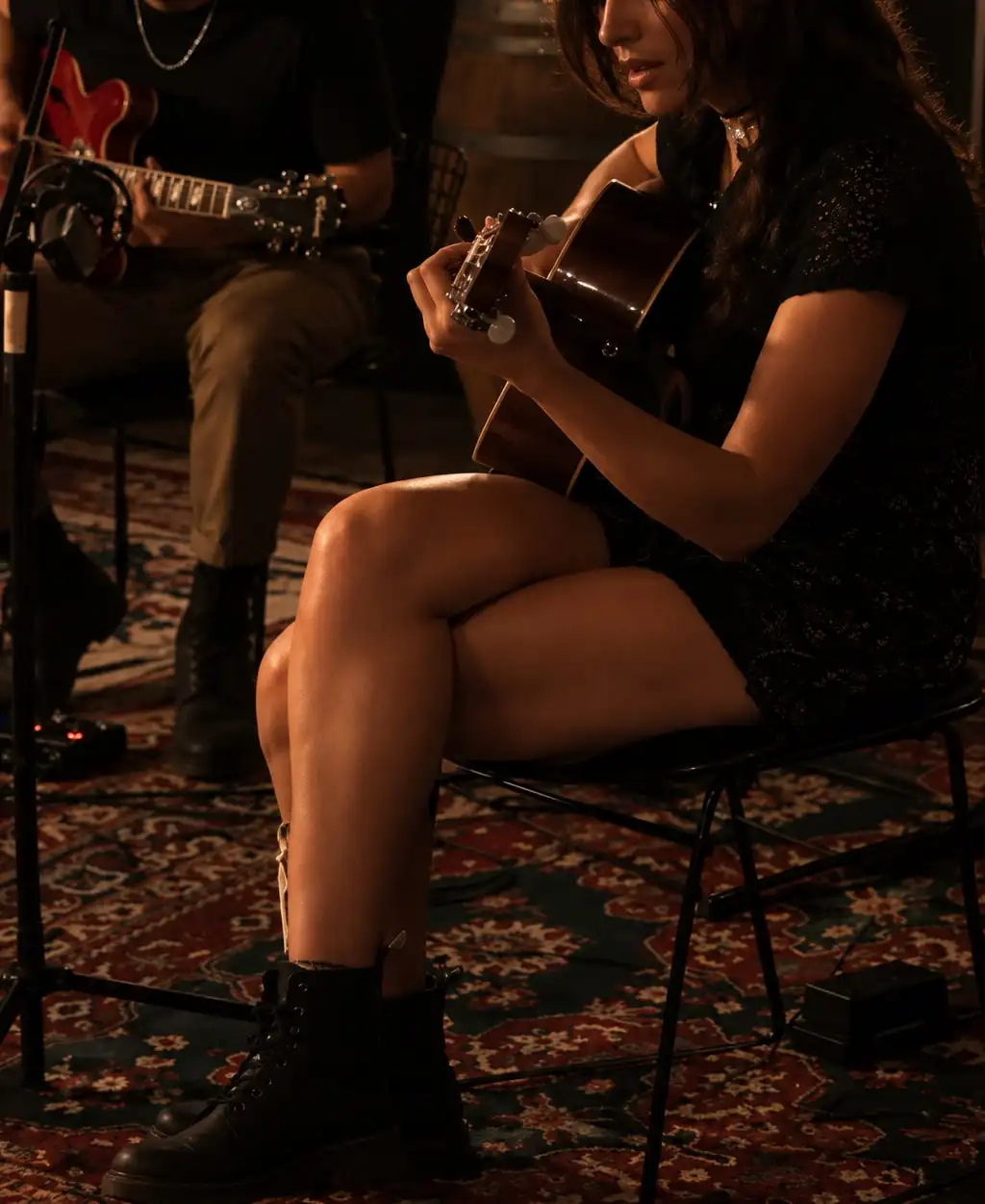 Una mujer toca la guitarra criolla sentada sobre una alfombra, con un guitarrista detrás.