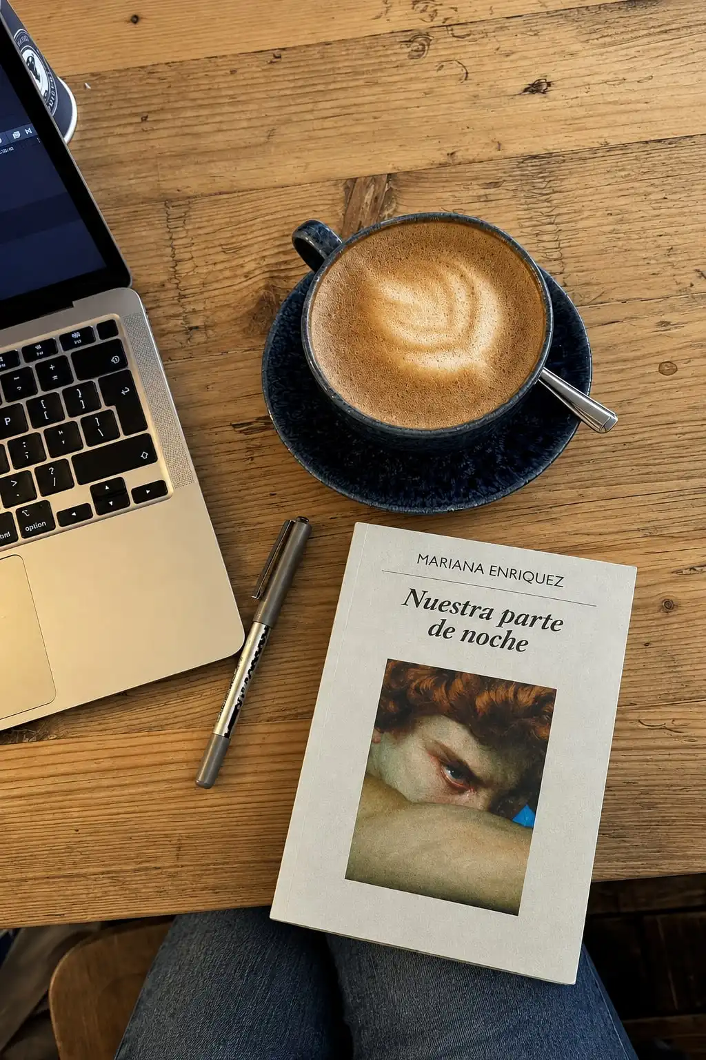 Una mesa de madera vista desde arriba: un café con leche en taza azul, una notebook, una lapicera y un libro.