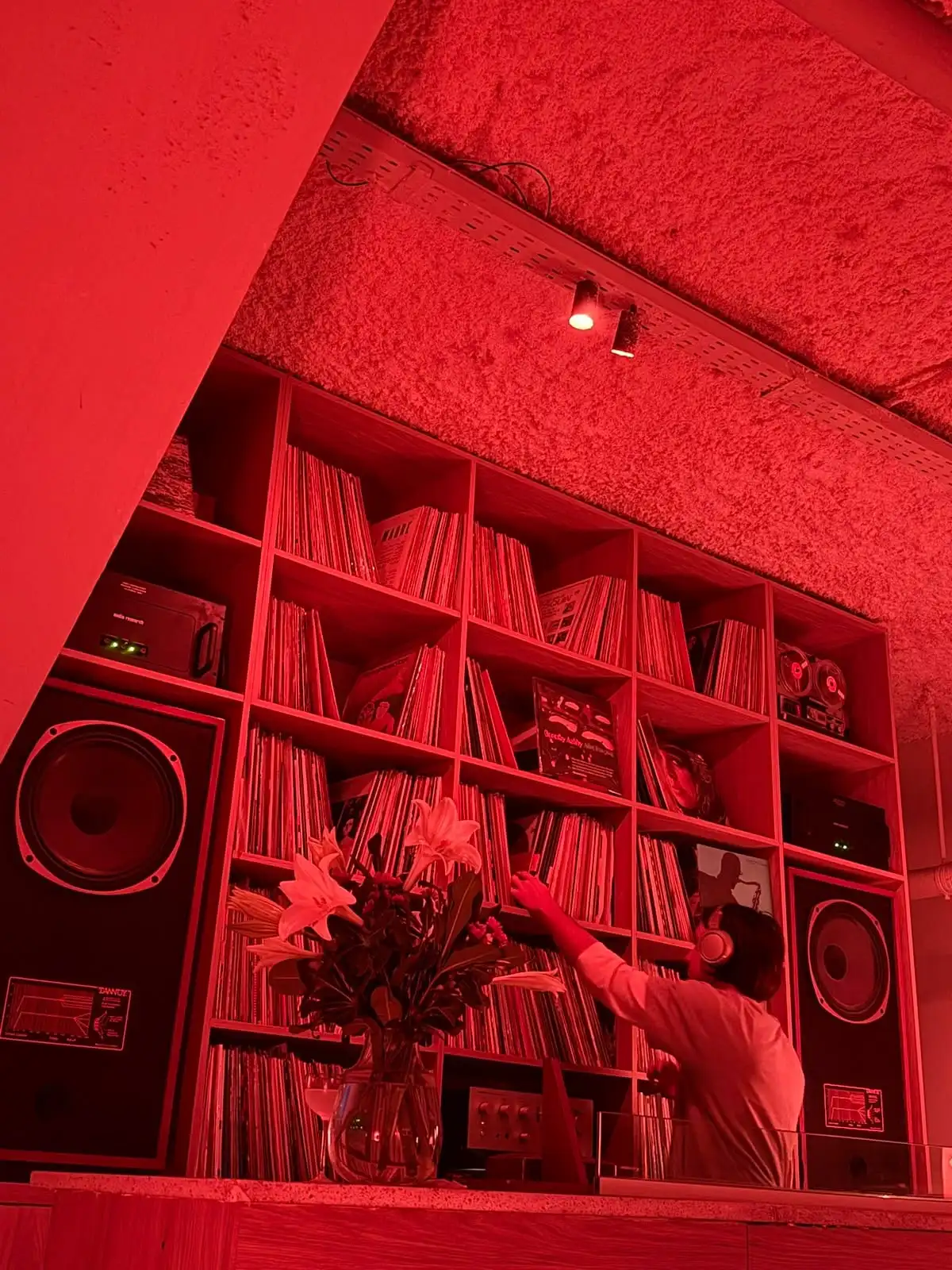 Una pared de estantes llenos de vinilos entre dos parlantes grandes, bajo luz roja; alguien con auriculares elige un disco.