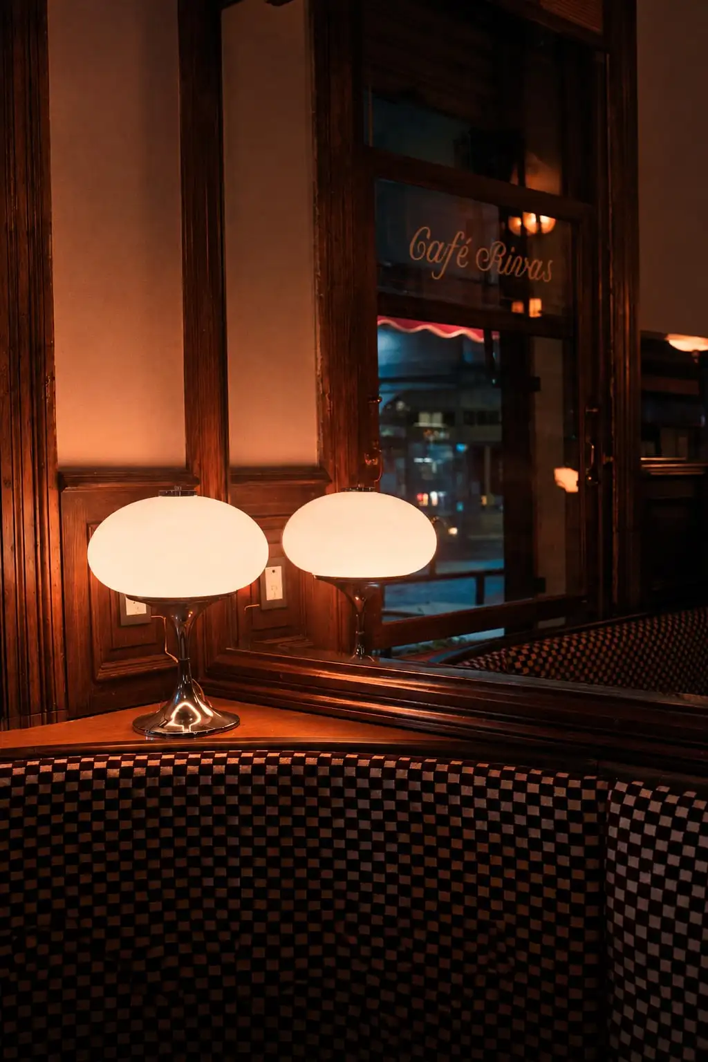 Rincón de un café antiguo de noche: dos lámparas de vidrio sobre la mesa, un sillón a cuadros y el nombre «Café Rivas» en la ventana.