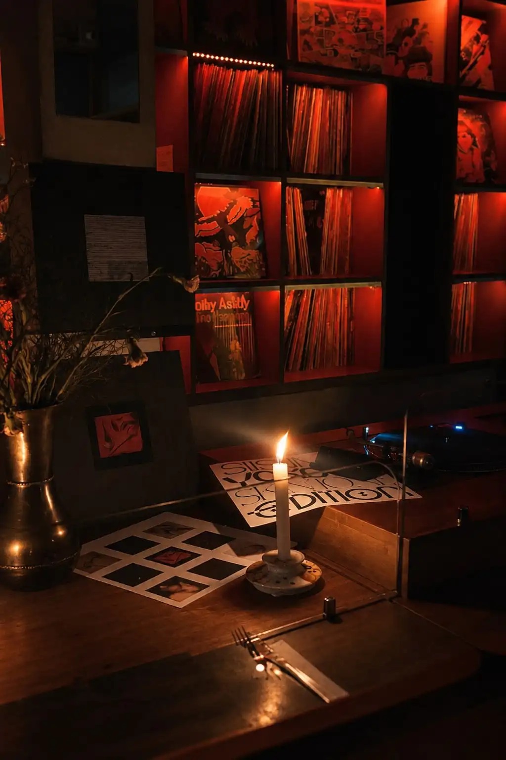 Una vela encendida sobre una mesa de madera, con estantes de vinilos iluminados de rojo y una bandeja de discos al fondo.