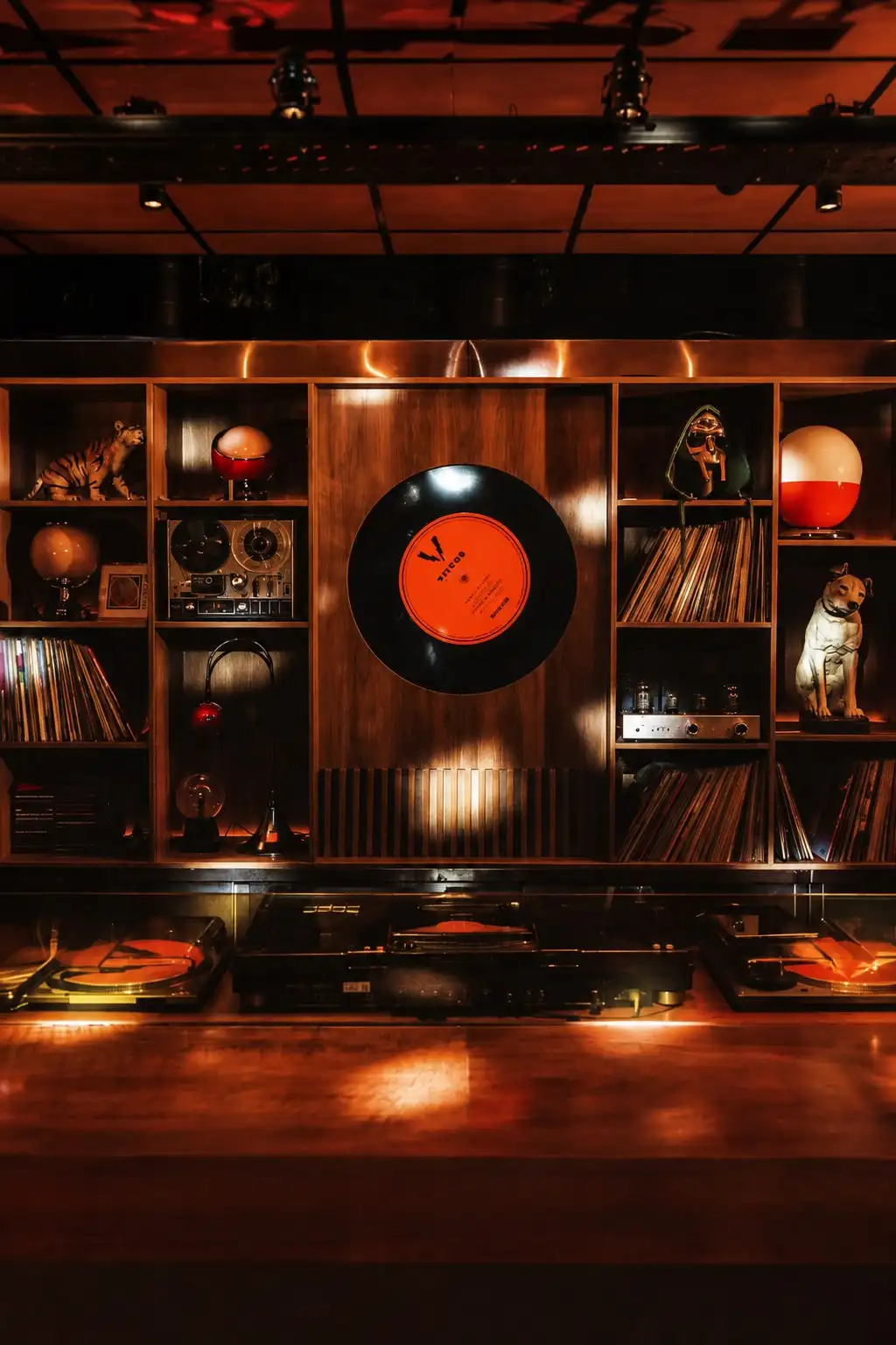 Mueble de madera con discos, un grabador de cinta abierta y objetos antiguos alrededor de un vinilo naranja colgado en el centro.