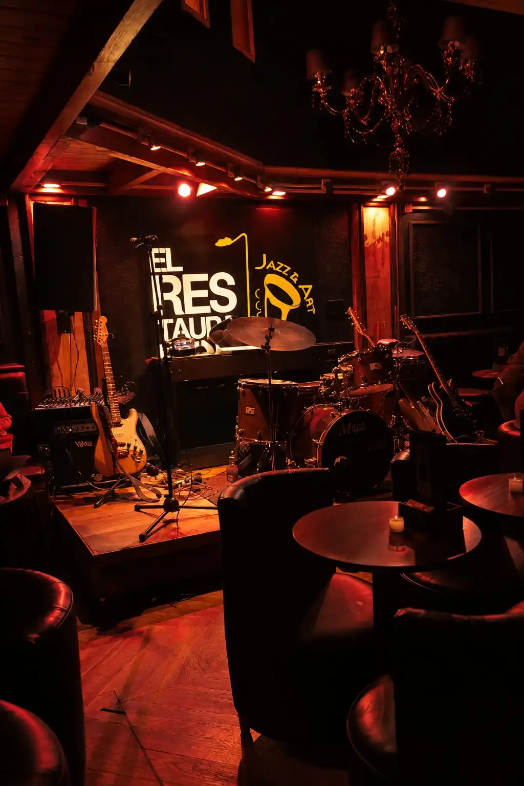 Escenario chico con batería, guitarras y un piano, y mesas redondas con velas adelante.