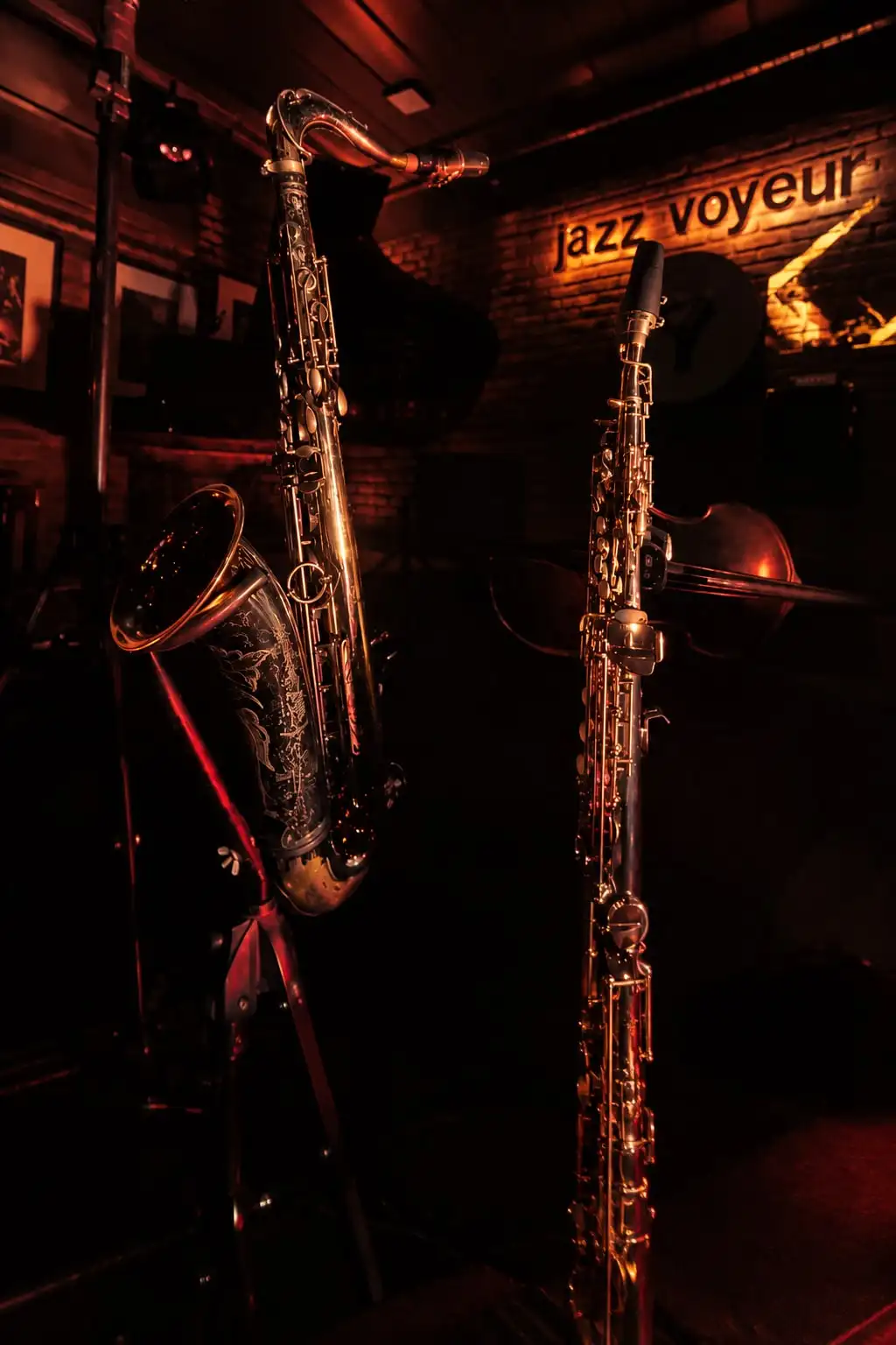 Dos saxofones en sus soportes sobre un escenario oscuro, con el cartel «Jazz Voyeur» en la pared de ladrillo.