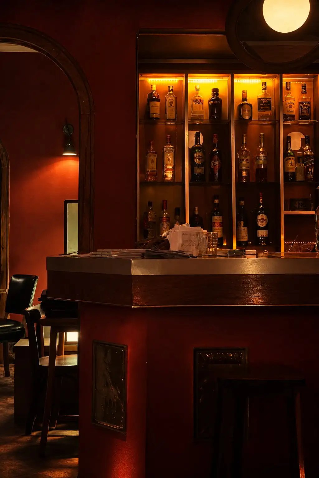 Barra de un bar de paredes rojas, con una vitrina de botellas iluminada al fondo.