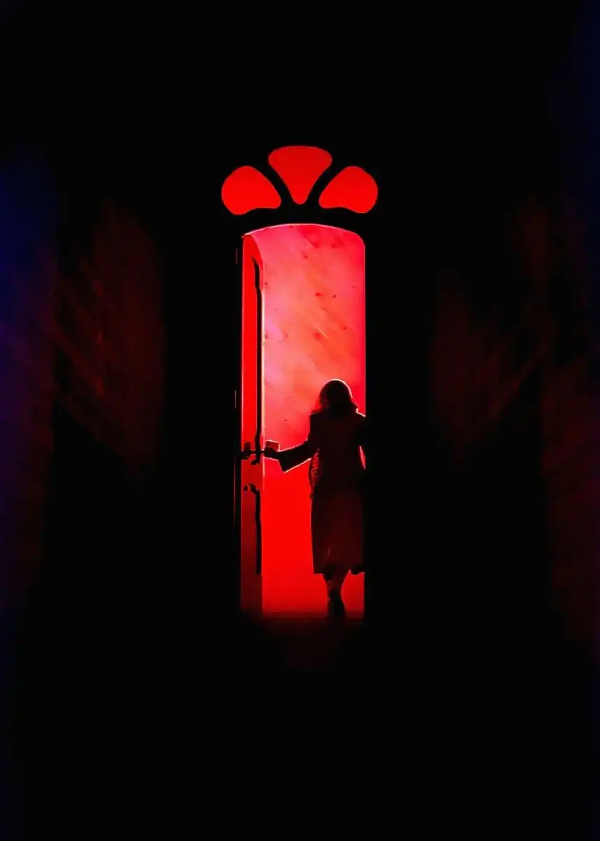 La silueta de una mujer abriendo una puerta alta iluminada de rojo, en un pasillo a oscuras.