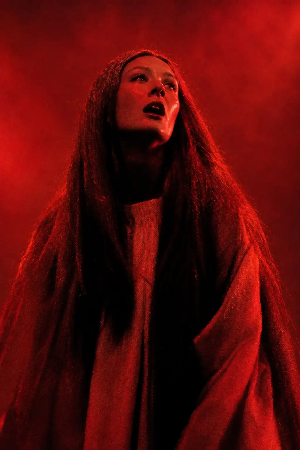 Una mujer de pelo largo mira hacia arriba con la boca entreabierta, envuelta en niebla y luz roja.