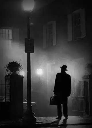 En blanco y negro, un hombre de sombrero y maletín parado bajo un farol, frente a una casa entre la niebla.