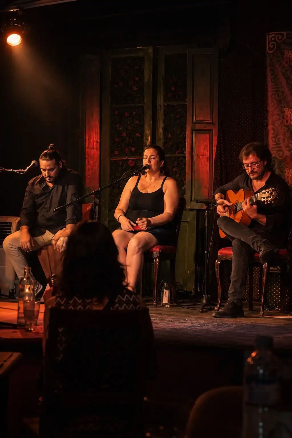 Un percusionista, una cantaora y un guitarrista sentados en un tablao, vistos desde el público.