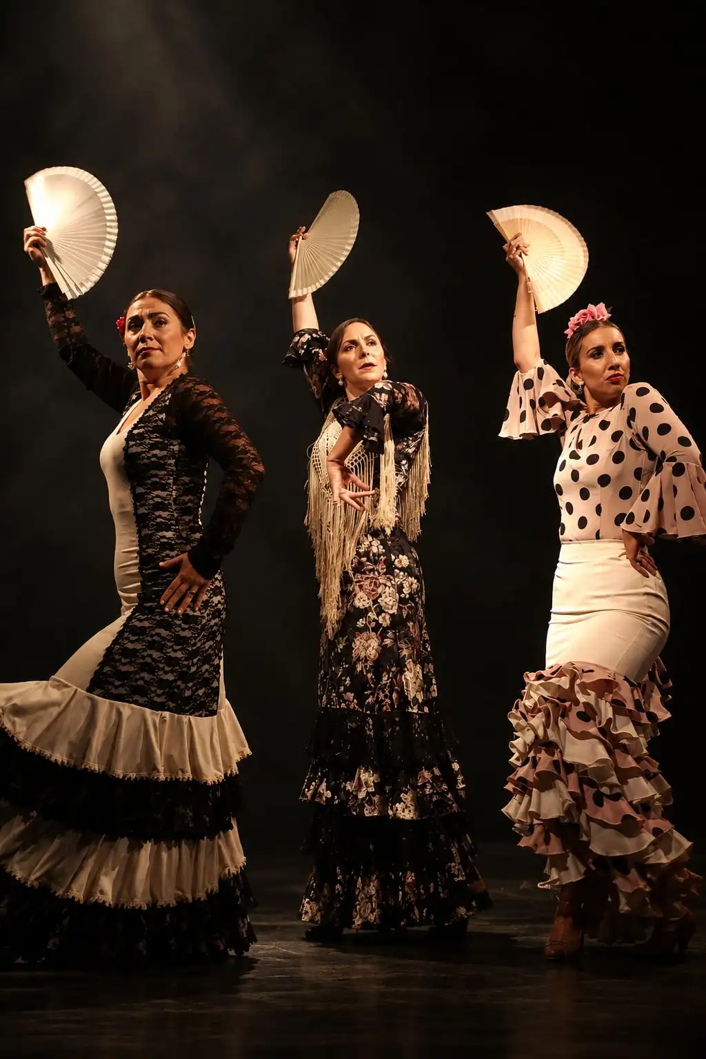 Tres bailaoras con vestidos de volados blancos y negros levantan sus abanicos en un escenario oscuro.