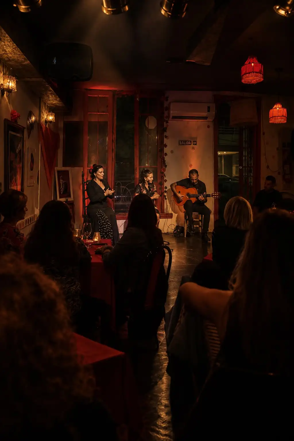 Un salón en penumbra con el público sentado de espaldas, mirando a los músicos al fondo bajo lámparas rojas.